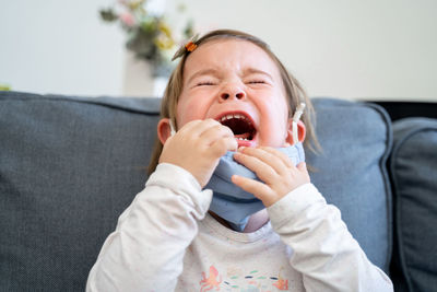 Close-up of crying toddler with mask sitting on sofa at home
