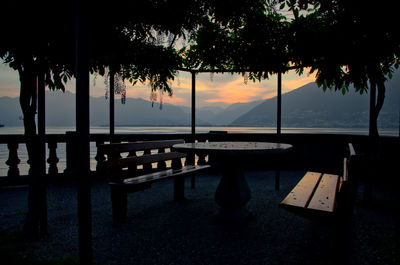 Silhouette trees by empty bench against sky during sunset