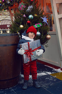 Christmas boy a child in warm clothes holds a gift and stay on a barrel next to a christmas tree