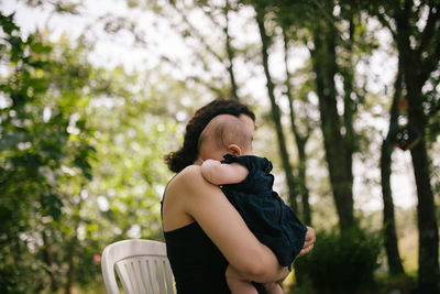 Side view of mother carrying baby girl against trees