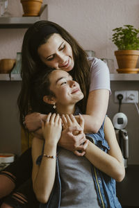 Smiling woman hugging female friend from behind while sitting in kitchen at home