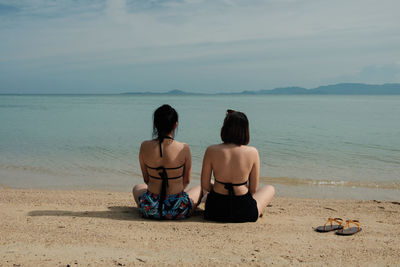 Rear view of female friends in bikini sitting on shore at beach against sky