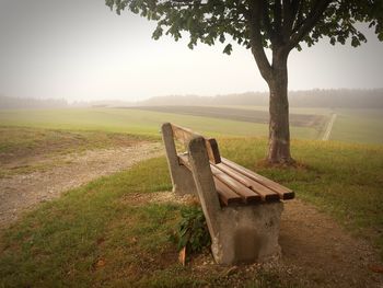 Bench on field by trees against sky