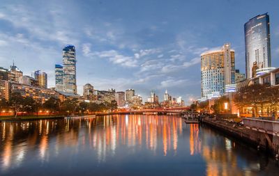 Illuminated buildings by river against sky in city