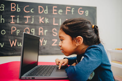 Side view of girl using laptop on table against blackboard