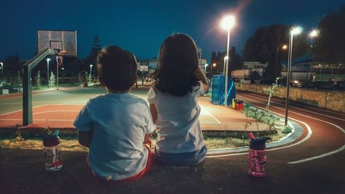 Rear view of kids sitting on retaining wall looking at illuminated basketball court