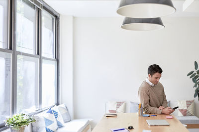 Businessman using mobile phone while sitting at table against wall in creative office