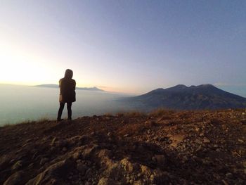 Rear view of person standing on mountain by sea against sky during sunset