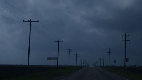 Empty road amidst electricity pylons on field against cloudy sky
