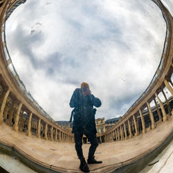 Rear view of man reflecting on mirror while standing at historic building against cloudy sky
