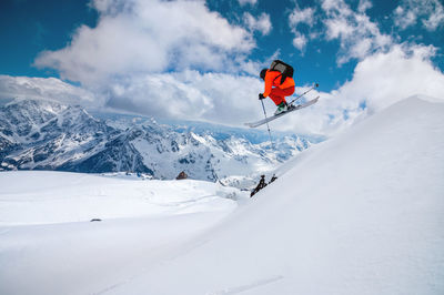 A freerider skier in an orange suit with a backpack froze in a jump flight over high snow-capped 