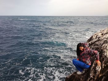 Full length of woman crouching on rock by sea against sky