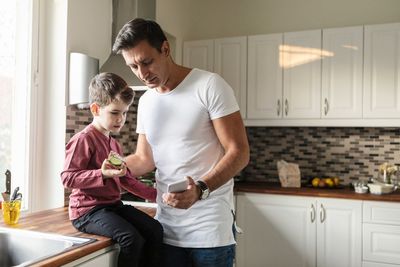 Father showing smart phone to son with food while standing by kitchen counter