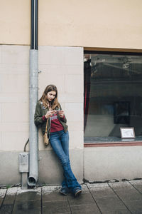 Full length of girl using smart phone while leaning on pipe against wall