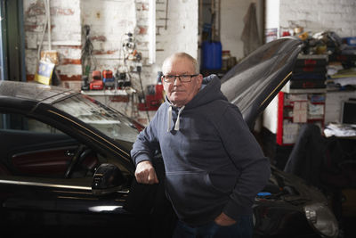 Confident male mechanic with hand in pocket leaning on car at workshop