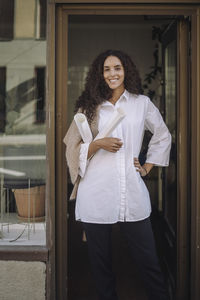 Portrait of smiling female architect standing with hand on hips at office doorway