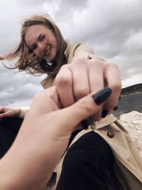 Cropped image of friend holding young woman hand against cloudy sky