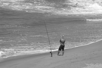 Rear view of shirtless man fishing while standing on shore at beach