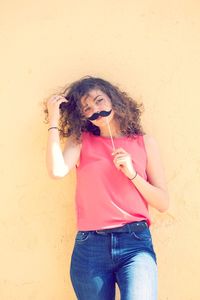 Portrait of young woman holding mustache prop while standing against wall