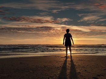 Rear view of boy standing at beach against sky during sunset