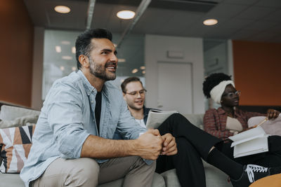 Happy mature businessman sitting by young colleagues sitting on sofa during conference meeting