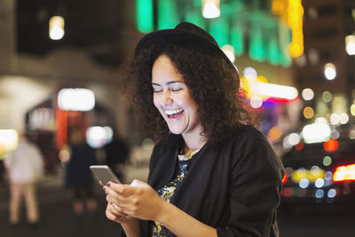 Young woman laughing while using smart phone in city at night