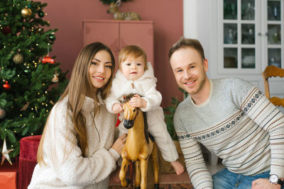 A small child of one year is sitting on a wooden horse near the christmas tree. his parents