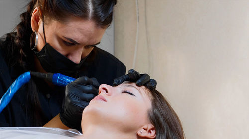 Close-up of beautician wearing mask coloring woman eyebrow in salon