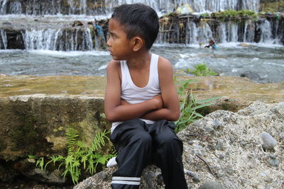 Wet boy looking away while sitting on rock by river