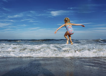 Full length of playful girl with arms outstretched jumping over shore against sky