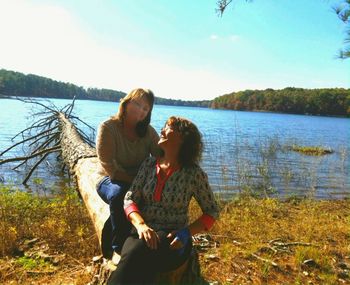 Friends sitting on grass by water against sky