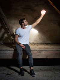 Full length of young man with hand raised sitting on wood indoors