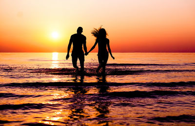 Silhouette couple running in sea against sky during sunset