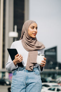 Content muslim female in headscarf walking with takeaway drink anfd tablet in the city while looking away