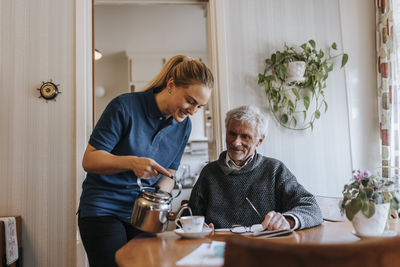 Smiling care assistant serving tea to senior man sitting at home