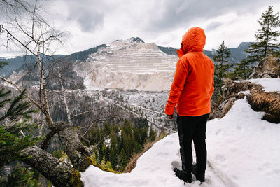 Rear view of person in orange jacket standing on snowcapped mountain during winter