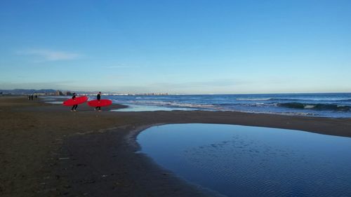 Man and woman carrying surfboards walking at sea shore against clear sky