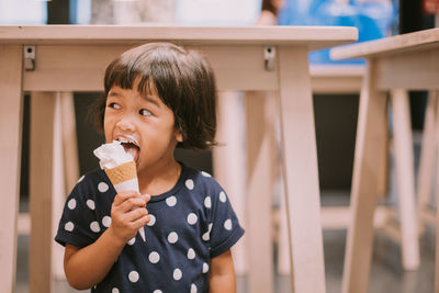 Cute girl eating ice cream while standing against tables