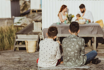 Rear view of male siblings sitting over blanket while family with baby girl in background