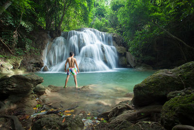 Rear view of shirtless man standing against waterfall at forest