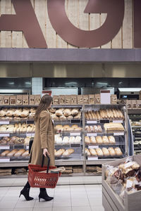Side view of woman walking with basket by breads in rack at supermarket