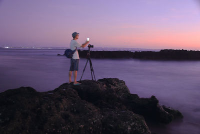 Man photographing on rock by sea against sky during sunset
