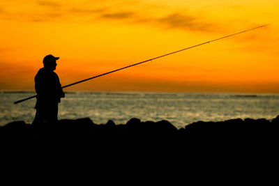 Silhouette man fishing in sea against sunset sky