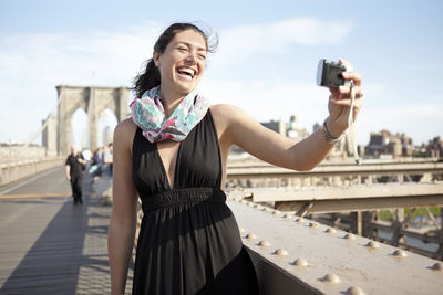 Woman in black dress taking selfie with brooklyn bridge in background