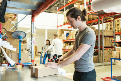 Manual worker packing box while coworkers discussing in distribution warehouse