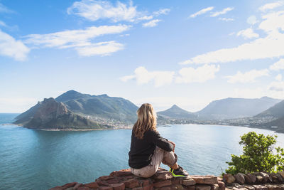 Side view of woman sitting on stone retaining wall while looking at hout bay against sky