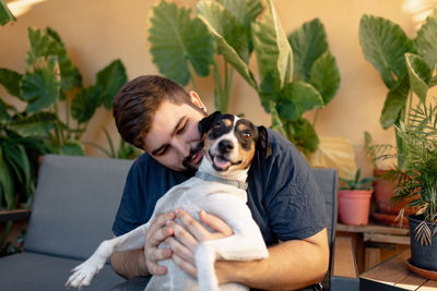 Young man smiling is holding up his dog while playing with his paws. the dog is looking at camera