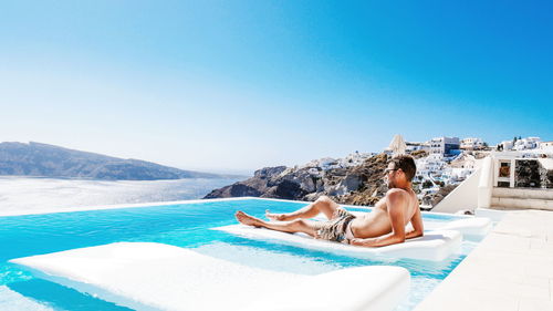 Shirtless man relaxing at infinity pool by sea against clear blue sky
