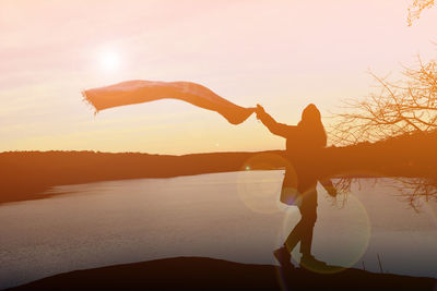 Full length of silhouette woman holding scarf by lake against clear sky during sunset