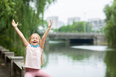 Cheerful girl with arms raised sitting on railing by canal